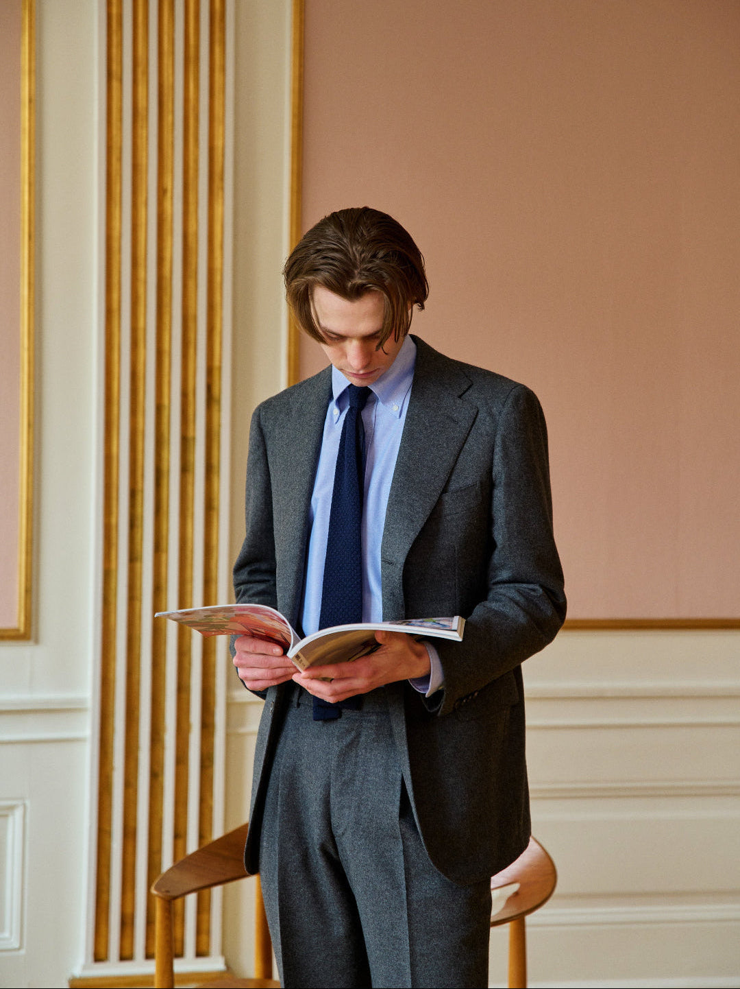 Man in a suit reading a book in an elegant room with wooden paneling.