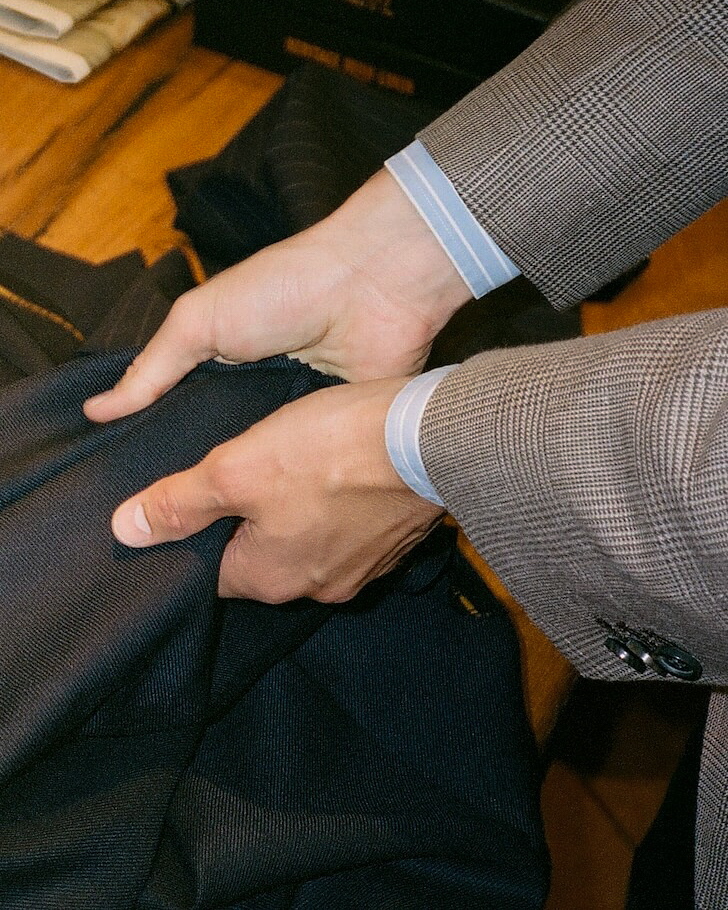 Close-up of a person's hands holding and folding a dark navy wool fabric, wearing a brown and gray checkered suit jacket with light blue dress shirts.