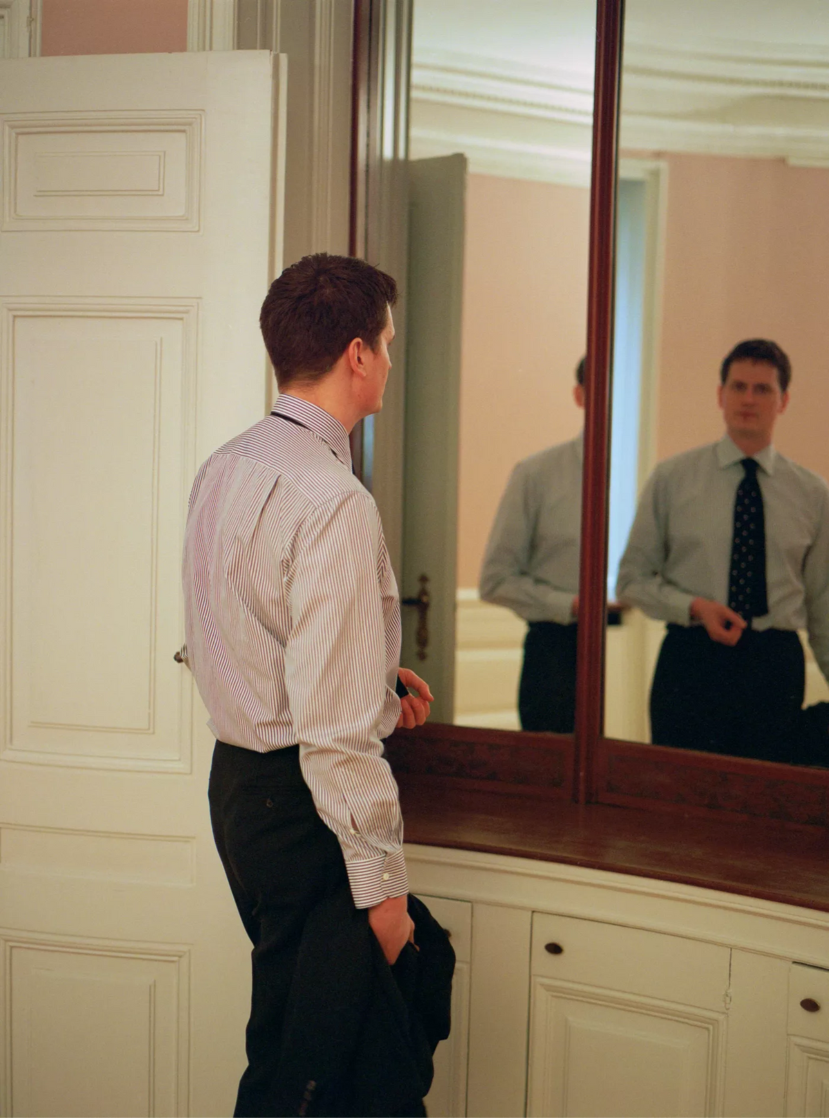 Man adjusting his tie in front of a large mirror in a room with white walls and wooden furniture.