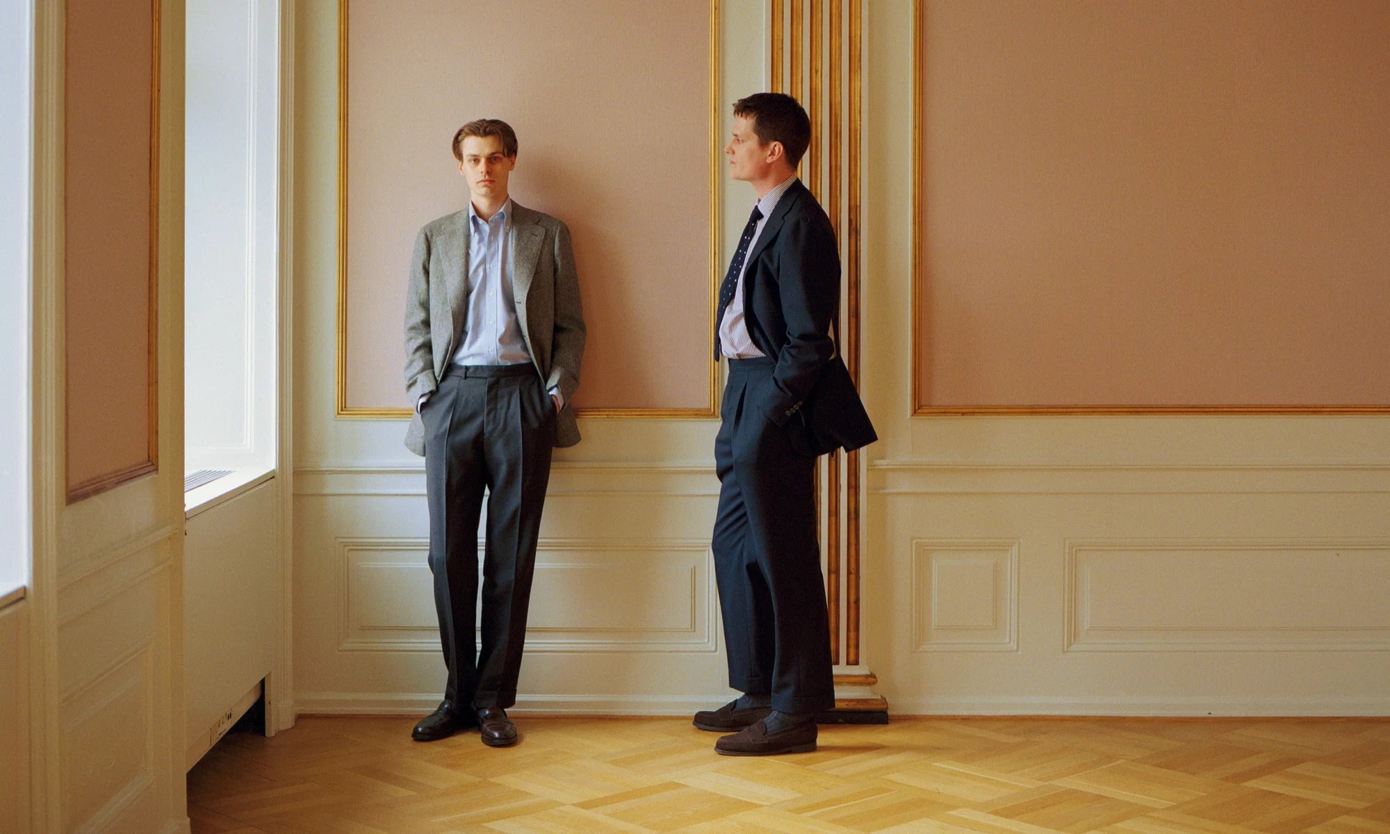 Two men in suits standing in a room with beige walls and wooden floor.