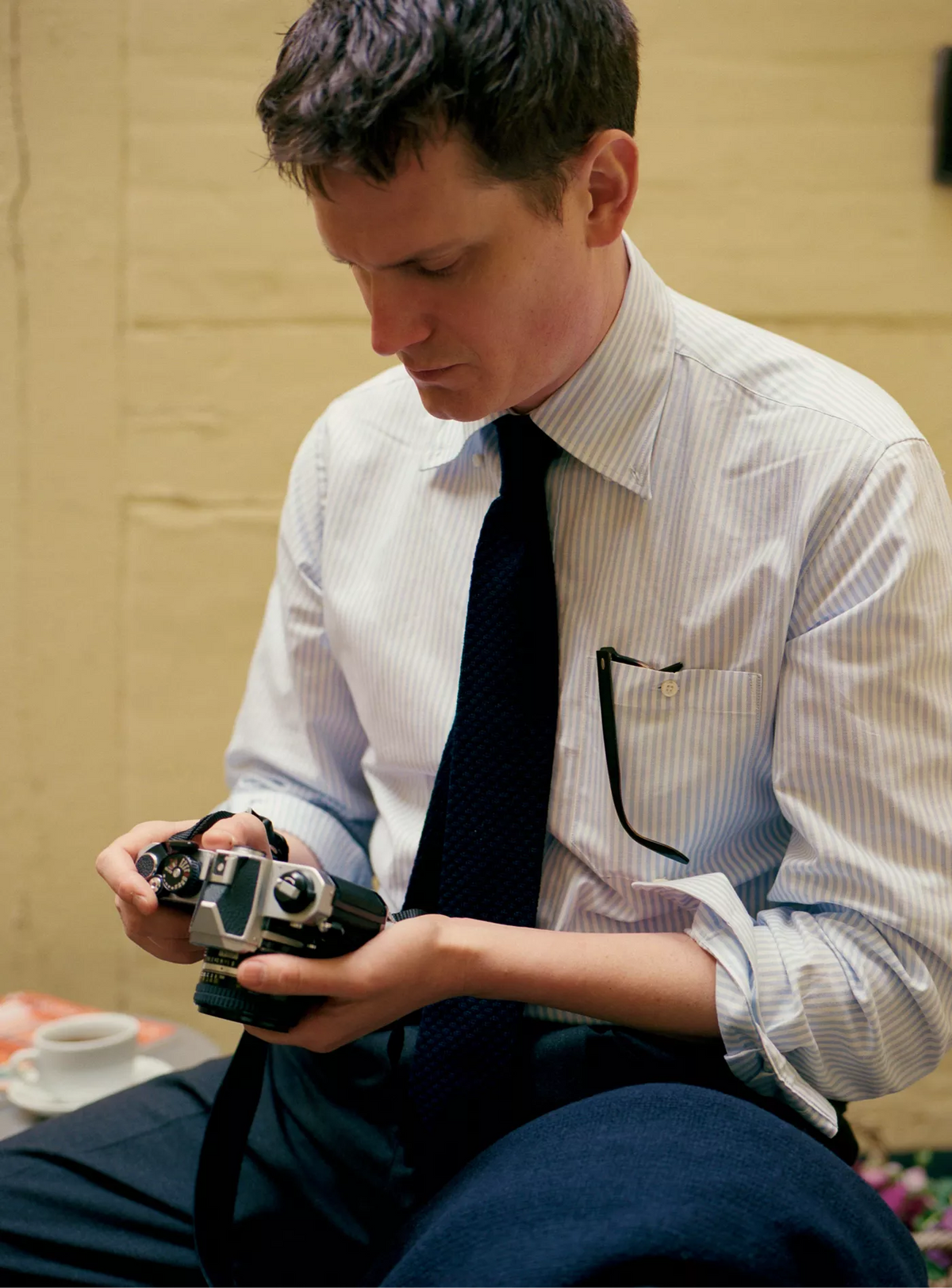 Man in a striped shirt and tie holding an old-fashioned camera against a yellow wall.